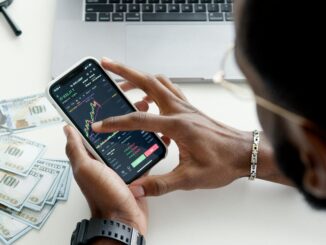 Person reviewing live trading chart on smartphone, laptop with stock charts open and cash on desk. Someone managing and earning money online from home